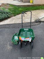 Photo showing two green fertilizer and grass seed spreaders, one large push type with wheels and one small handheld type on pavement outdoors.