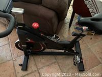 Side view showing the black frame, red flywheel, red adjustment knob, and pedal with foot strap.