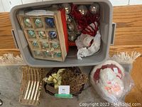 Overhead view of two plastic boxes containing various vintage glass Christmas ornaments, a wrapped Santa Claus figure, a gold-tone metal sleigh, and a brown wicker basket with faux greenery and ribbon bow.