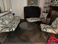 Metal framed 3-seat couch, two single chairs, large round glass coffee table on patterned carpet in indoor setting