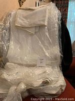 Five white linen table cloths, folded and wrapped in clear plastic, photographed on a chair indoors.