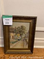 Framed painting by T.H. Wilkinson showing a village street with cottages, flowers, and a figure walking, signed at bottom left.