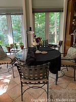 Set of round glass patio table with four metal chairs around it, cushions on chairs, set shown indoors in sunroom with tile floor.