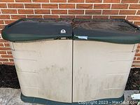 Photo of large Rubbermaid outdoor storage bin with beige body and dark green hinged lid, dirty exterior, placed against a brick wall.