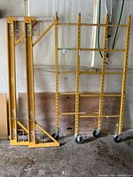 Yellow metal scaffolding components: two rectangular tower frames with caster wheels and two triangular A-frame support legs leaning against wall.
