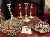 Photo shows four silver-colored metal candle holders and three silver-colored metal trivets with ornate floral and leaf cut-out designs arranged on a red surface.