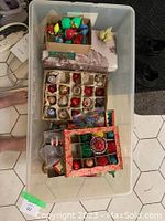 Plastic string lights, multiple boxes of glass ornaments and ceramic bird visible inside white storage bin