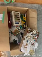 Large cardboard box open on floor showing assorted wrapped glass bird ornaments, a string of mini lights, and other small decorations