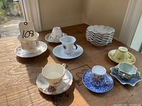 Overview of nine assorted tea cups and saucers displayed on a wooden table under natural light, showing various colors and styles.
