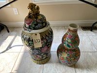 Two large Chinese decorative vases on tile floor; one black floral vase with gold dog lid and one double-gourd shaped floral vase
