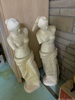 Photo showing two tall cream-colored porcelain Aphrodite bust statues with missing arms, placed side by side on a floor against a wall.
