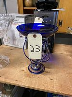 Photo showing deep cobalt blue glass display or candle holder on wooden surface next to other household items. The piece has a round shallow bowl atop a decorative base with three teardrop-shaped supports.