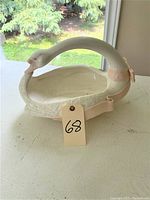 Overall view of white ceramic swan basket with a curved neck handle and pink accents sitting on white surface with outdoor background.
