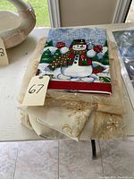 Four folded table linens stacked on a table, with the top one featuring a colorful snowman Christmas design. Others include lace and plain linens.