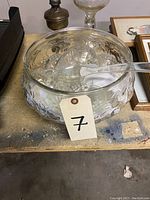 Clear glass punch bowl with embossed floral design, holding 10 clear glass cups and a clear glass ladle inside it, placed on a table alongside other items.