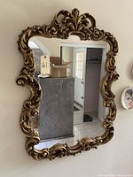 Full frontal photo of large wall mirror with ornate gold carved frame hanging on a wall showing its intricate scrolling and floral detailing.