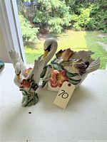 Two white ceramic swan figurines with floral bases on display near window, showing overall size and detail.