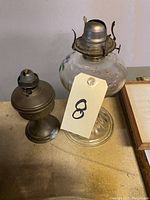Photo of two vintage oil lamps on a beige surface, one glass with floral decoration and one metal, tagged with number 8.
