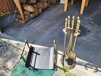 Photo showing one brass andiron wood holder and a brass stand holding four fireplace tools on a concrete floor.