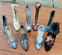 Photo of nine decorative ceramic shoe figurines arranged on a wooden table, showing various styles including boots and high heels with assorted colors and decorative details.