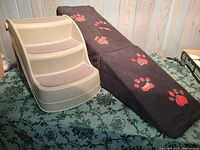Side view of beige plastic dog stairs with carpeted steps and adjacent black foam ramp covered in red paw print fabric.