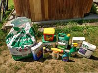 Wide photo showing all gardening supplies spread on grass including the bag of peat moss, assorted fertilizers, weed killer, and sprayers.
