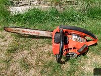 Photo of an orange Homelite XL-2 gas chainsaw with black handles and metal guide bar lying on grass.