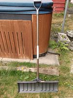 Full view of the snow shovel leaning against an outdoor wooden structure with metal handle and wide plastic blade.
