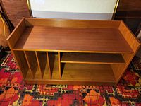 Front view of teak veneer stereo bench showing removable vertical record dividers on left and open shelving on right under flat top surface with raised edges.