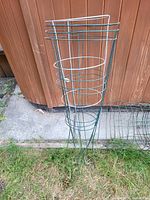 Stack of metal tomato plant cages leaning against a wooden fence showing the different heights and conditions of the cages, some with visible rust and wear.