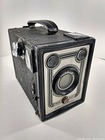 Angled photo showing front and side of vintage Vredeborch Vrede Box camera with textured exterior, lens, two windows, and top handle.