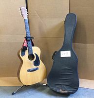 Full view of Anjo acoustic guitar on metal folding stand with red strap, natural wood finish and black pickguard