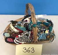 Woven basket filled with assorted costume jewellery featuring multiple colors and styles of beaded necklaces, bracelets, and a watch.