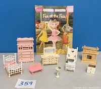 Full set of eight doll house furniture pieces arranged in front of a colorful 'Fun at the Fair' sticker book, with visible details of shapes and colors.