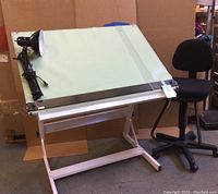 Drafting table with green slanted top, attached straightedge, black adjustable chair, and clamp-on lamp seen from angle