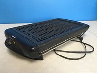 Black T-Fal electric raclette grill with temperature dial and power cord, slotted grill surface, sitting on a white table with blue background.
