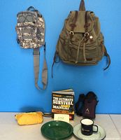 Photo showing front view of two backpacks, survival manual, tin plates, tin mug, child's raincoat, and water bottle in case.