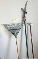 Full view of rake, cultivator, and hedge trimmer standing upright against a plain white wall showing all three tools together.
