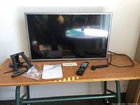 Front view of the LG TV with wall mount parts, remote control, owner's manual, and power cable arranged on wooden table.