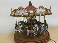 Front view of the electric Christmas carousel showing white and black horses mounted on gold poles, decorated with painted winter scenes and gold trim on the top canopy.