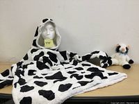 Photo of cow print fleece hoodie displayed on a mannequin head and spread flat on surface with the cow plush toy next to it.