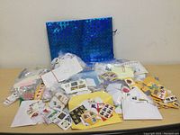 Photo showing multiple envelopes, plastic bags, and loose stamps spread out on a table including a large blue iridescent gift bag in the background.