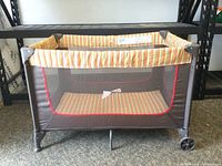 Playpen shown from the front and side, positioned on the floor below a metal shelving unit.