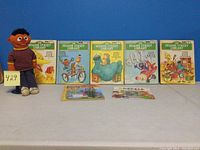 Five Sesame Street Library hardcover books with colorful character covers, standing upright next to a plush Ernie doll wearing a striped sweater and light pants, plus two coloring/activity books laid flat in front
