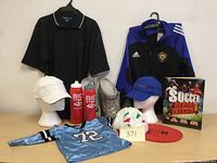 Full lot showing all items including clothing, book, caps, water bottles, Crocs, frisbee, and soccer ball arranged on table and hanging