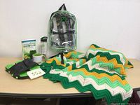 Shows all items in the lot together on a table: Afghan blanket, clear backpack, flip flops, water bottle, book, and mug.