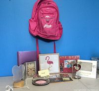Front view photograph showing the pink Roots backpack hanging on a blue wall behind several picture frames, CDs, and a small lamp arranged on a table.