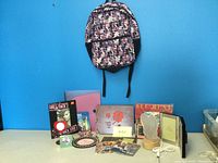 Photo showing the front of a multicolor patterned Roots backpack, a photo album, various picture frames, CDs, and a small night light arranged on the floor against a blue wall.