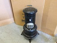 Black cast iron Valore oil stove showing ornate perforated vent design and three curved legs on floor against cardboard backdrop.