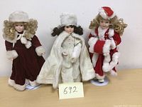 Front view of three Victorian Christmas collector dolls dressed in velvet and faux fur winter outfits, displayed against a plain white background with tag number 692.